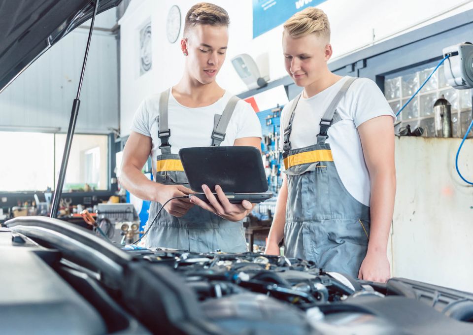 Zwei junge Automechaniker untersuchen ein Fahrzeug mit einem Laptop